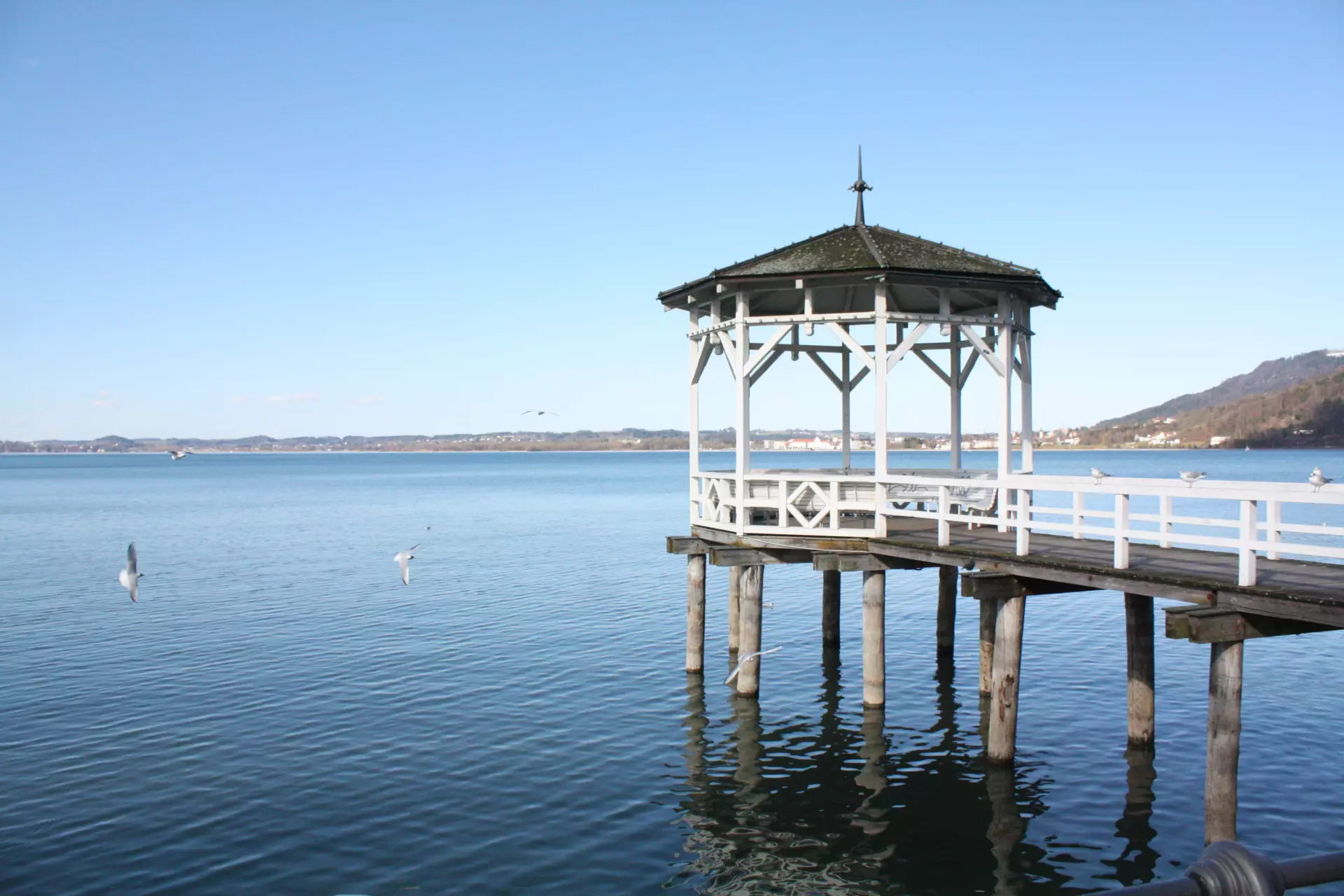 Weißer Steg mit Pavillon, der ins ruhige Wasser des Bodensees bei Bregenz führt, bei klarem Himmel und entfernten Hügeln im Hintergrund