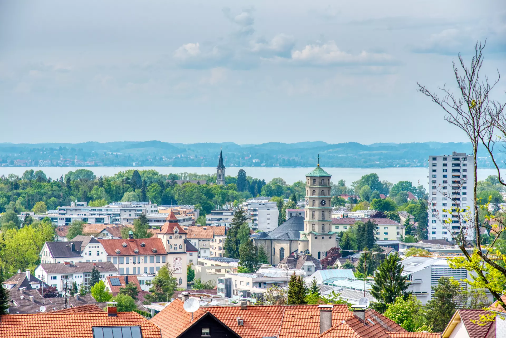 Blick auf die Stadt Bregenz mit Kirchtürmen, Wohnhäusern, Bäumen und dem Bodensee im Hintergrund unter bewölktem Himmel