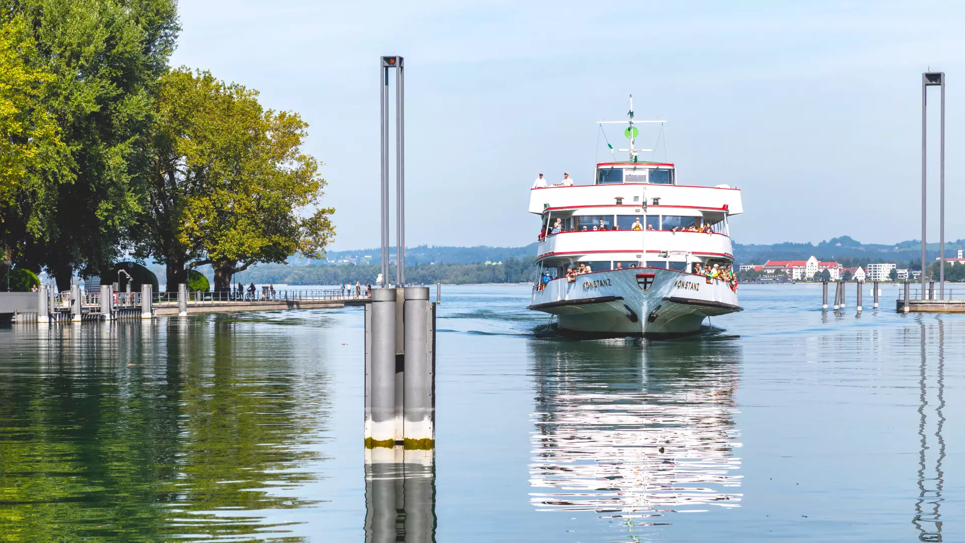 Passagierschiff fährt auf dem Bodensee in Bregenz entlang einer Uferpromenade mit Bäumen bei klarem Himmel