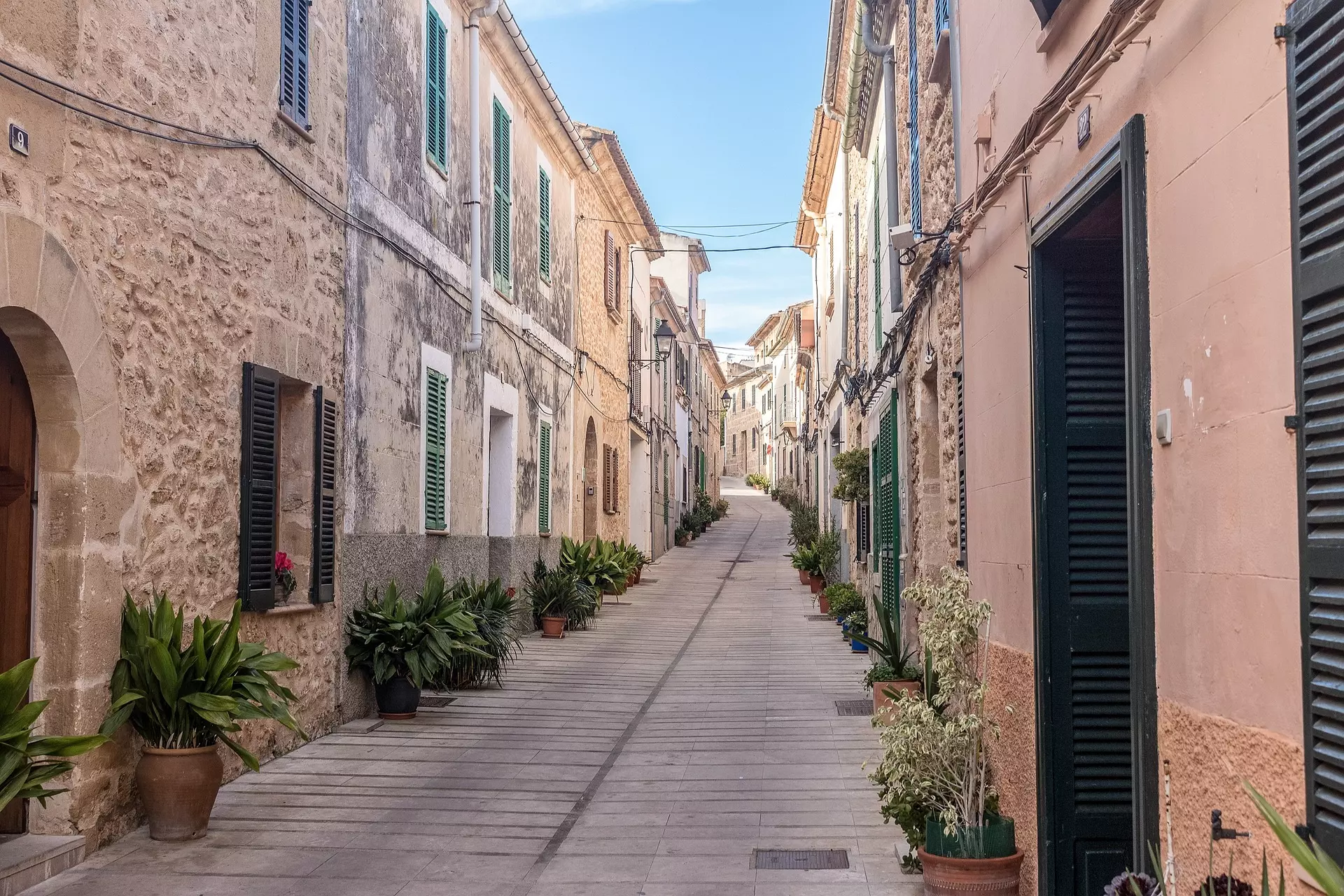 Schmale Gasse mit gepflastertem Boden, steinernen Häusern und Pflanzen in Töpfen auf Mallorca unter blauem Himmel