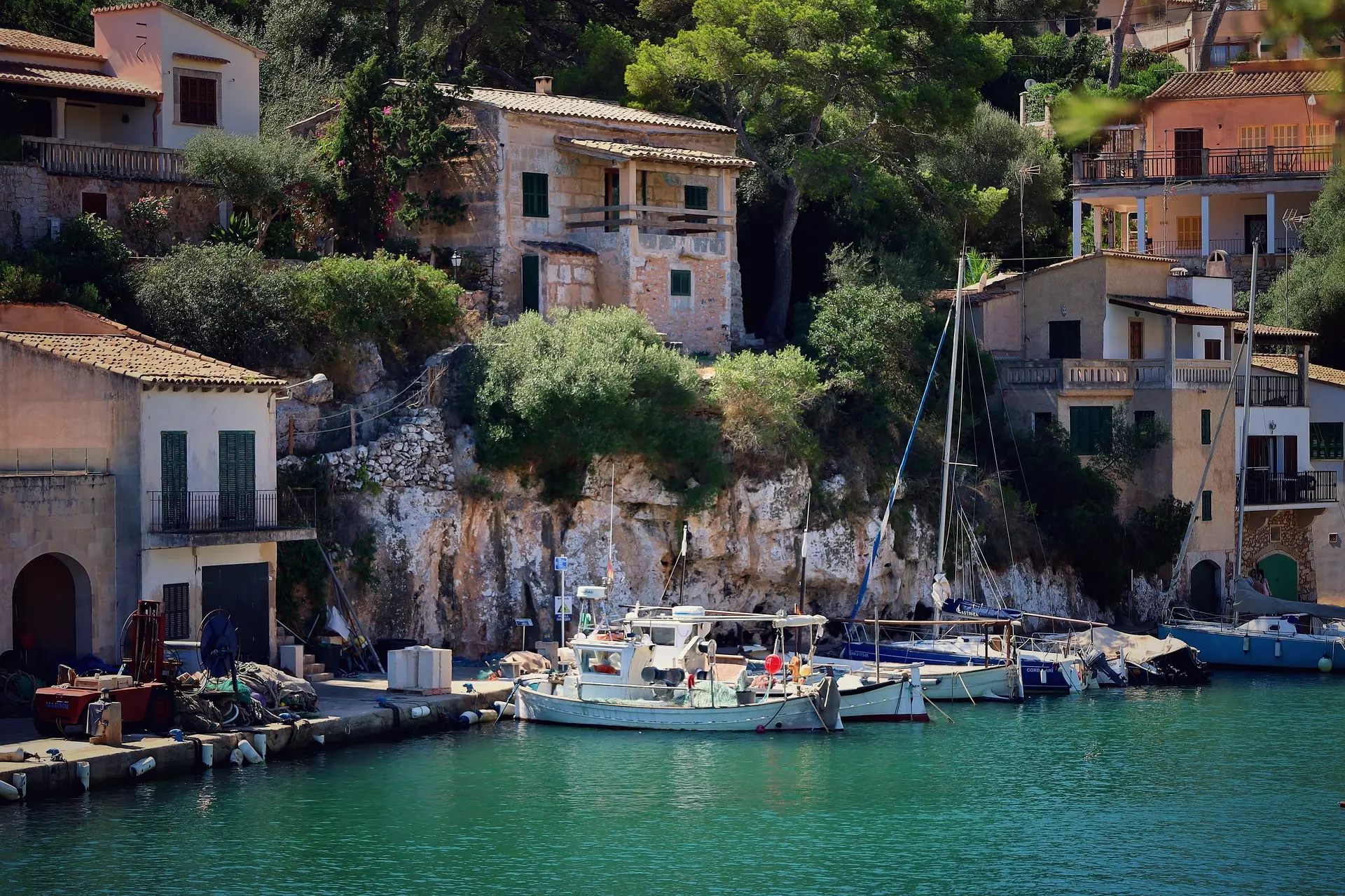 Boote am Hafen vor traditionellen Häusern und Felsen mit grüner Vegetation in Mallorca bei Sonnenschein