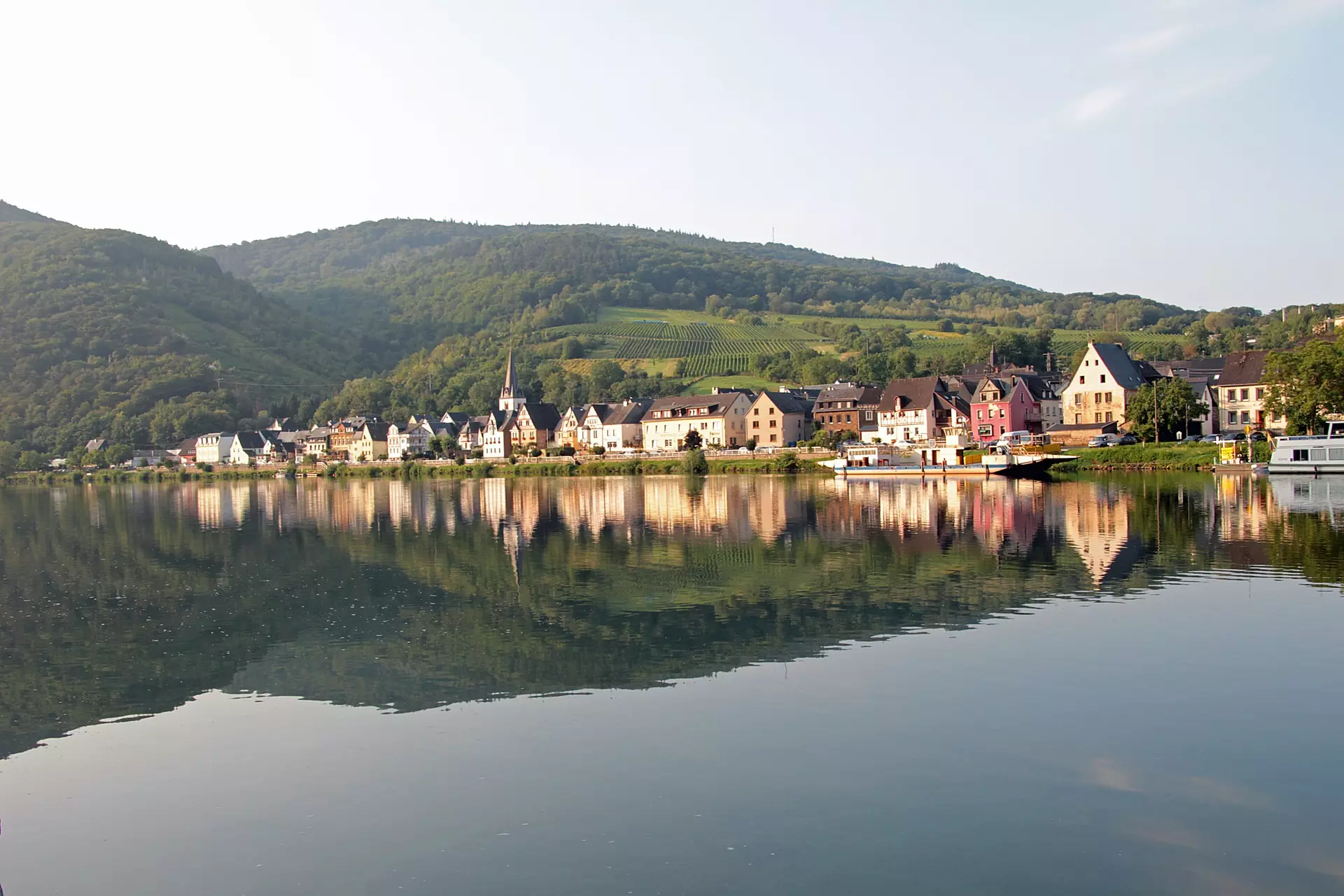 Blick auf die Moselregion mit ihren Weinbergen Moselregion mit Weinbergen, kleinen Häusern am Flussufer und ruhiger Wasserreflexion bei klarem Himmel