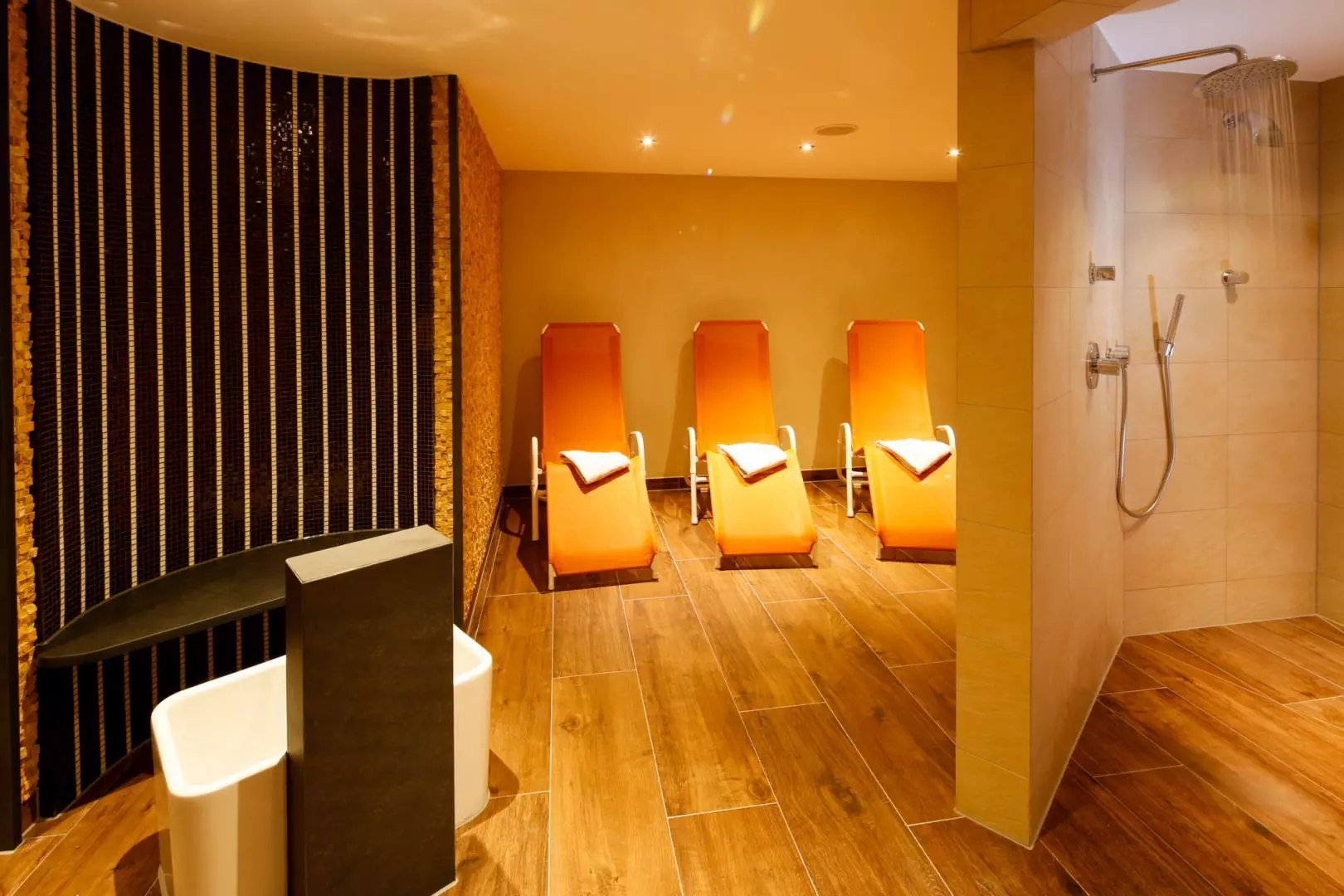 Entspannungsbereich mit drei orangen Liegestühlen, Holzfußboden und zwei Duschstationen im Hotel Messmer in Bregenz