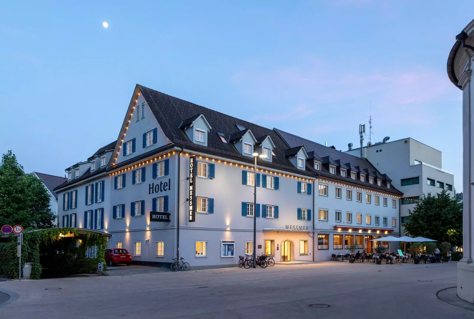Hotel Messmer in Bregenz bei Abenddämmerung mit beleuchtetem Gebäude und Sitzbereich im Freien am Straßenrand
