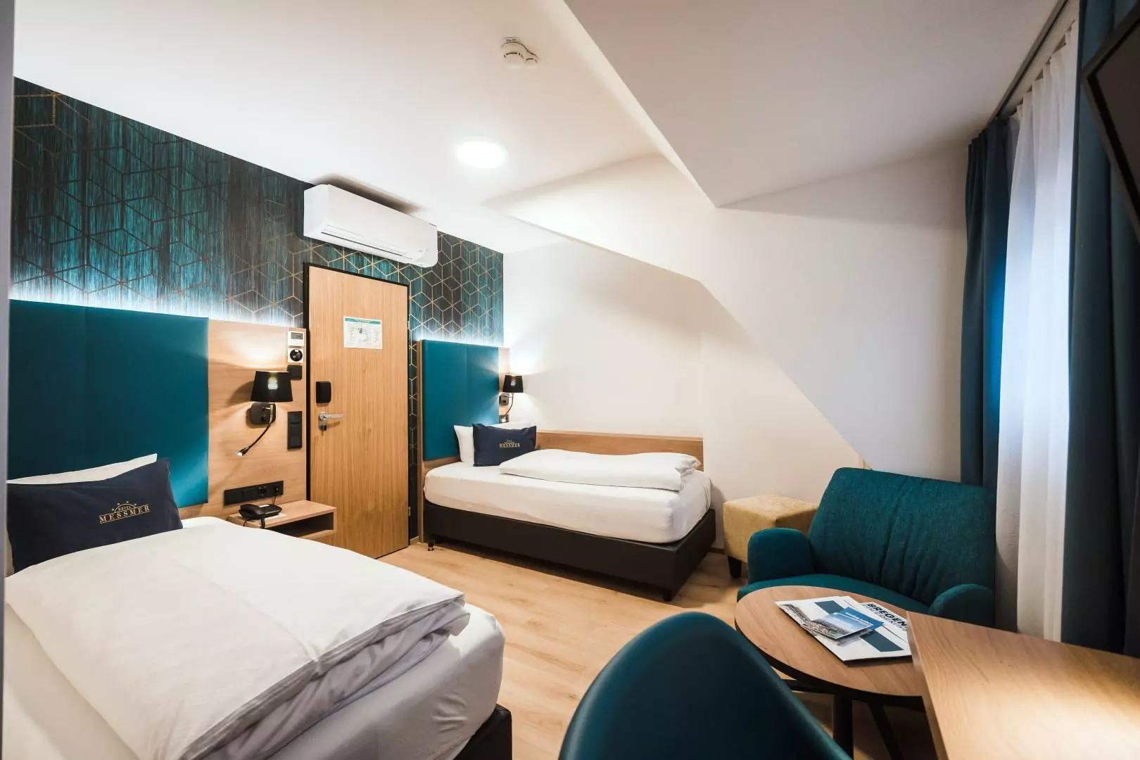 Modernes Hotelzimmer im Hotel Messmer Bregenz mit zwei Einzelbetten, Schreibtisch, Sitzecke und stilvoller Wandbeleuchtung
