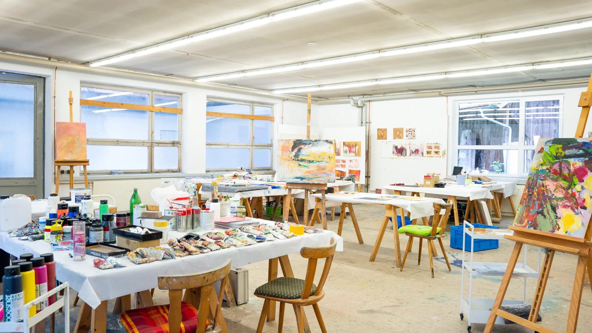 Kunstakademie Allgäu Helles Künstleratelier mit Staffeleien, Tischen voller Malutensilien, Farben, Pinseln und teilweise begonnenen Gemälden in der Kunstakademie Allgäu