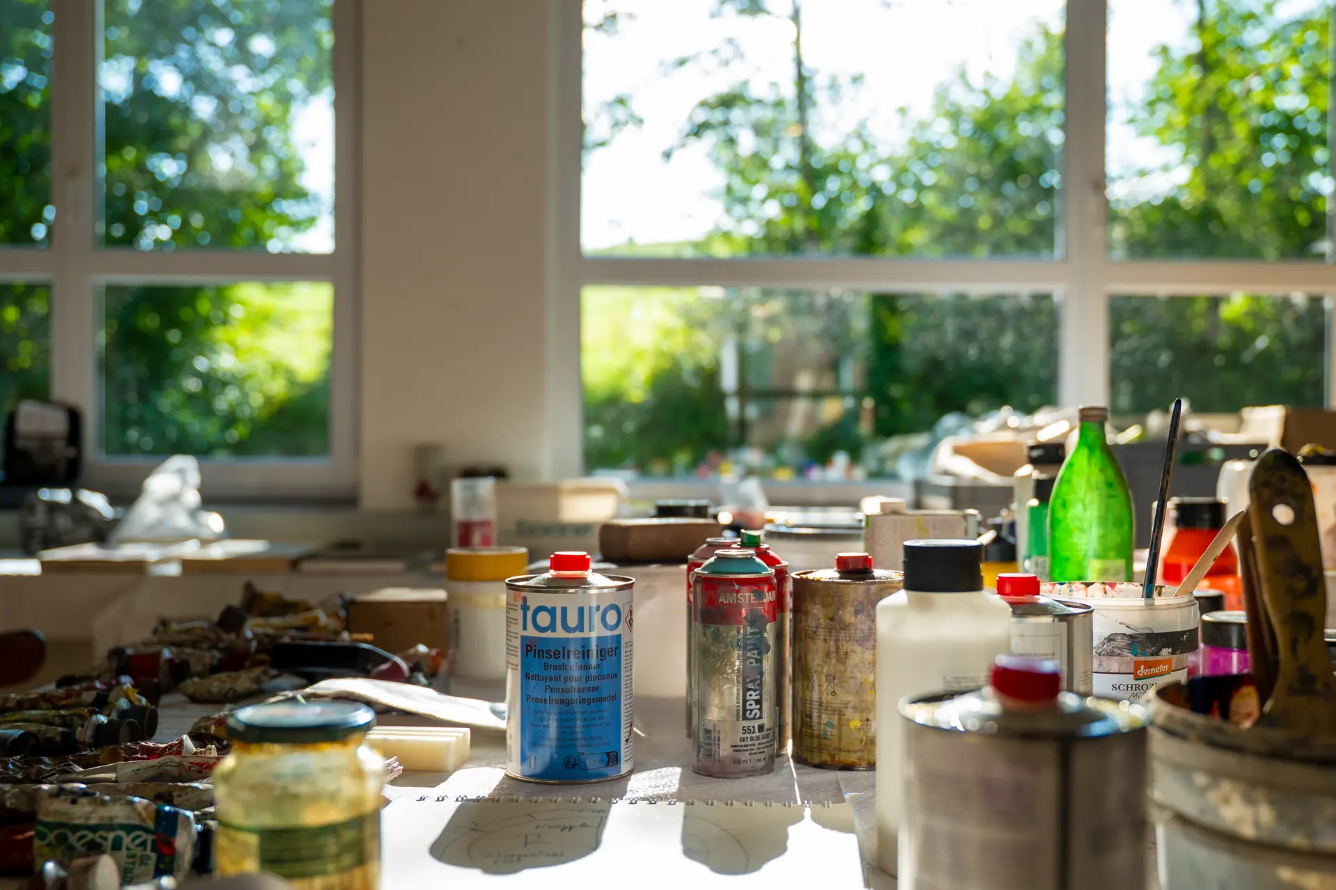 Kunstakademie Allgäu Atelier mit verschiedenen Farbdosen, Sprühdosen und Malutensilien vor großen Fenstern mit Blick auf grüne Bäume im Kunstakademie Allgäu in Allgäu