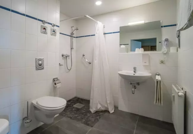 Barrierefreies Badezimmer mit bodengleicher Dusche, weißem Vorhang, Toilette, Waschbecken und großem Spiegel im Hotel Birschel-Mühle Hattingen