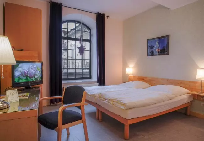 Einfach eingerichtetes Hotelzimmer im Hotel Birschel-Mühle mit Doppelbett, Fernseher, Holzstuhl und großem Fenster in Hattingen