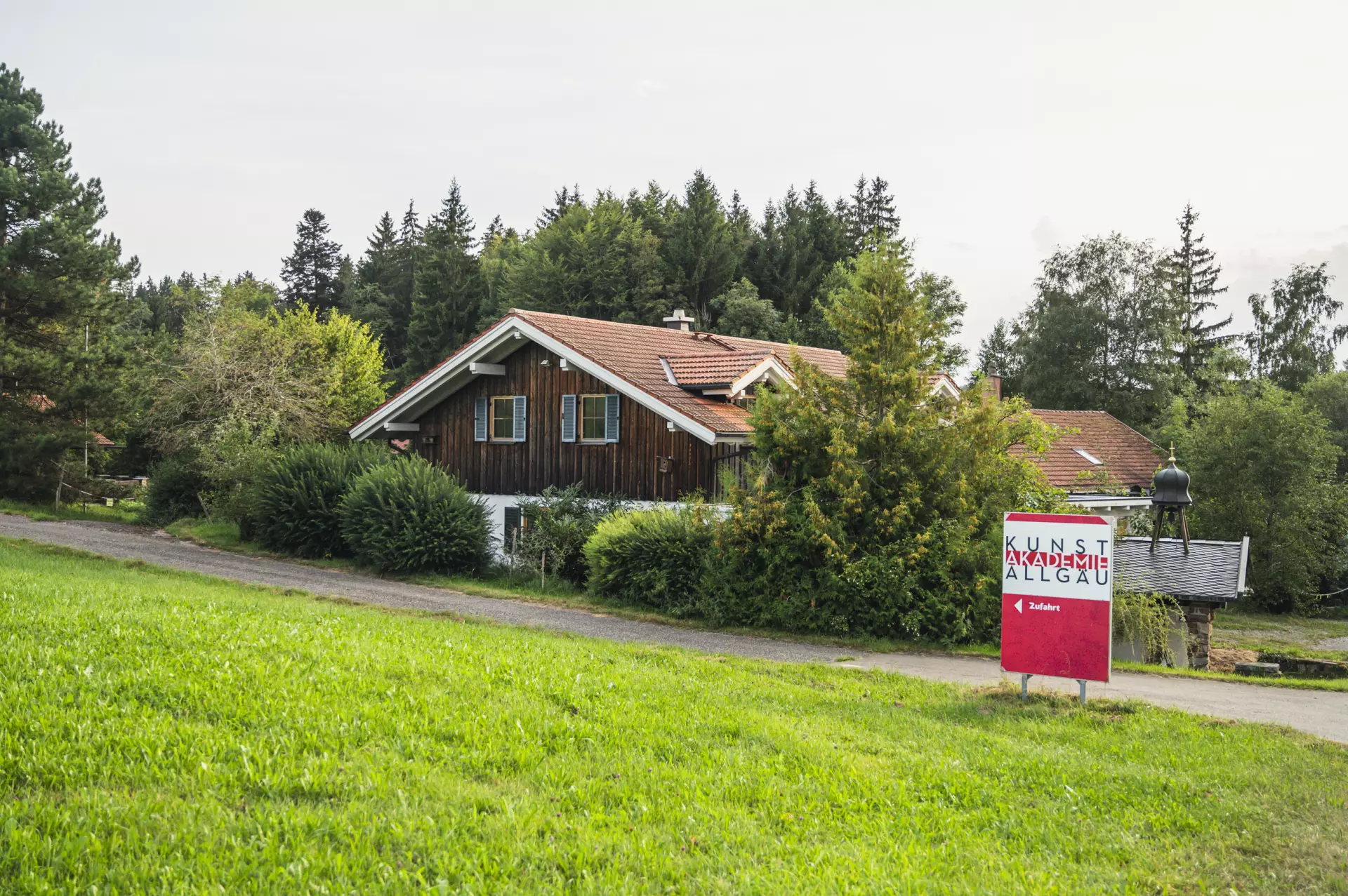 Holzhaus mit roten Ziegeldach umgeben von Bäumen und Wiesen vor Wald, daneben rotes Schild mit Aufschrift "Kunstakademie Allgäu" und Hinweis zum Atelier-Eingang
