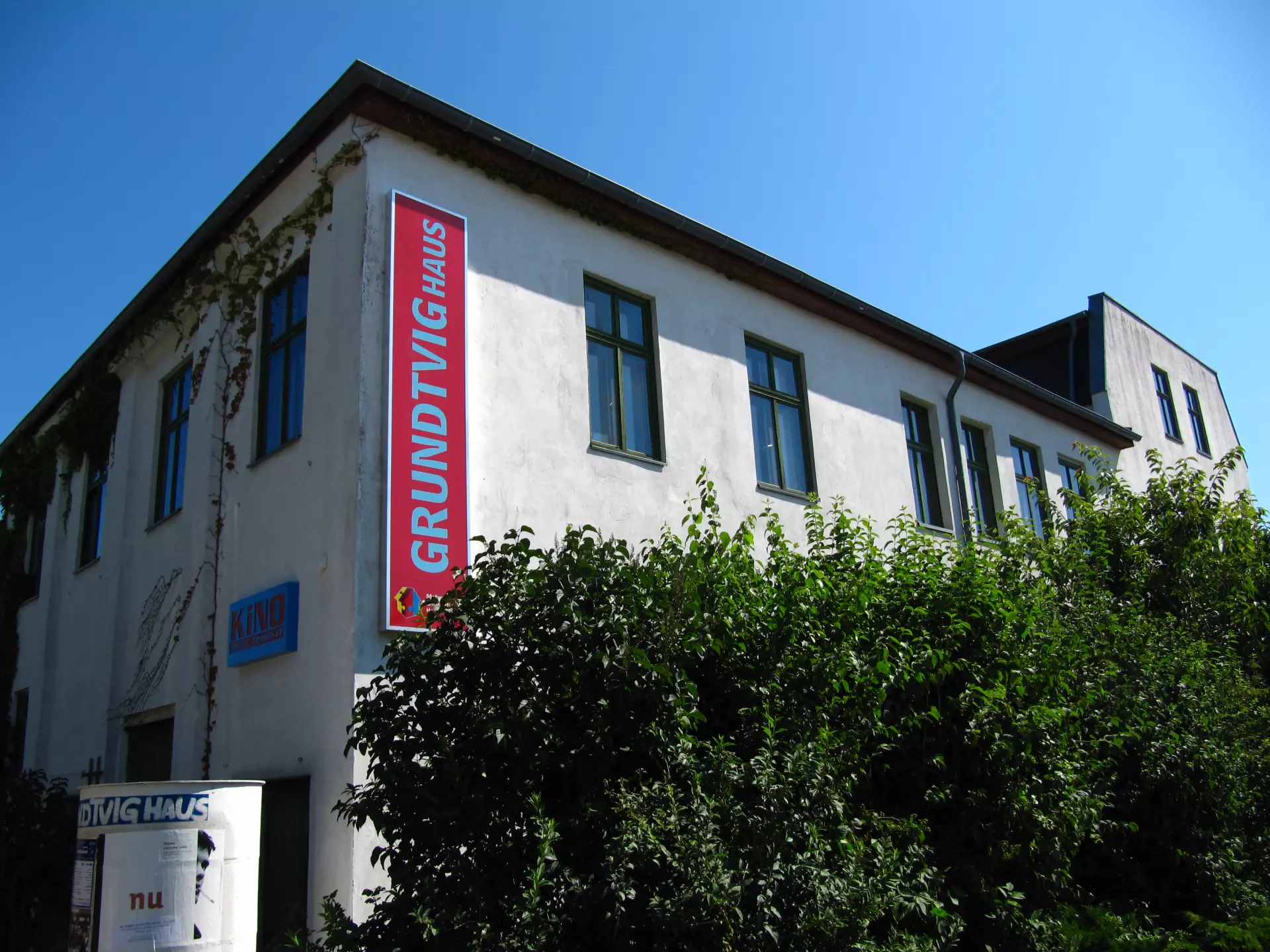 Atelier Grundvigthhaus Ruegen 2009 Sebastian(05) Mehrstöckiges weißes Gebäude mit grünen Fenstern und rotem Schild "Grundtvighaus" vor blauem Himmel und grünen Büschen in Rügen