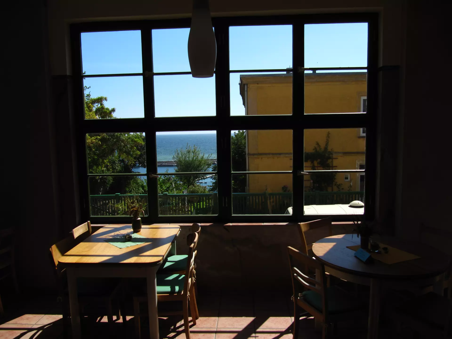 Atelier Grundvigthhaus Ruegen 2009 Sebastian(28) Sitzbereich im Atelier Grundtvighaus auf Rügen mit Holztischen, Stühlen und Blick durch ein Fenster auf das Meer und ein gelbes Gebäude