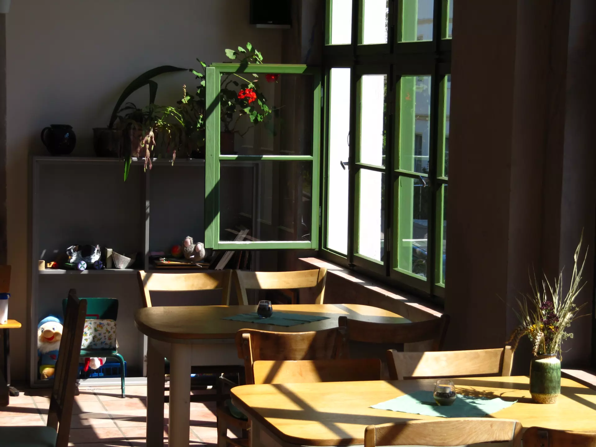 Atelier Grundvigthhaus Ruegen unterer Wintergarten 2009 Sebastian(31) Sonnenbeschienene Tische und Stühle im Atelier Grundtvighaus auf Rügen mit offenen grünen Fenstern und Pflanzen im Innenraum
