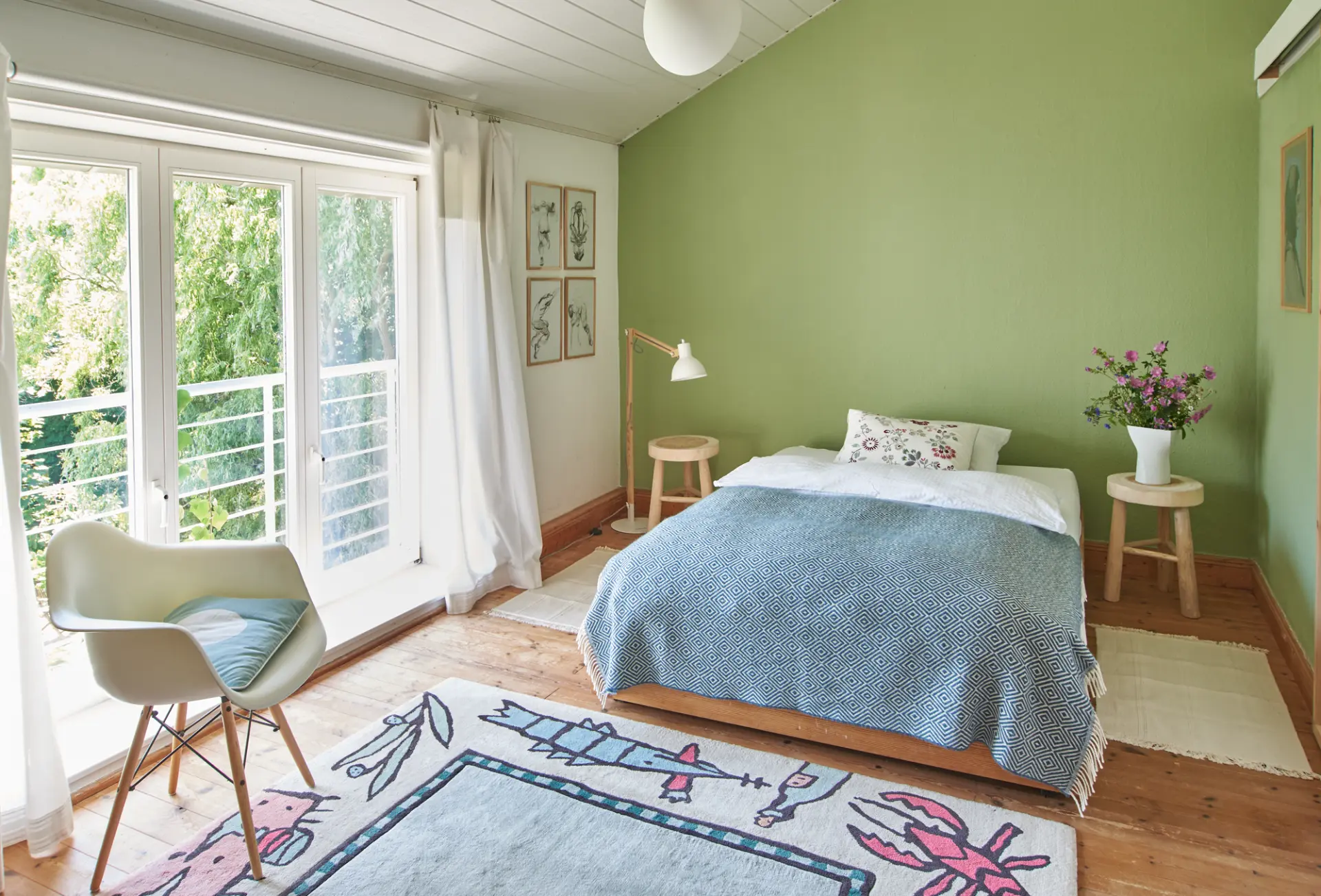 Schlafzimmer Werkgut Schlafzimmer mit grün gestrichener Wand, großem Fenster, Holzstuhl, Bett mit blauem Überwurf und buntem Teppich im Naturpark Aukrug Werkgut
