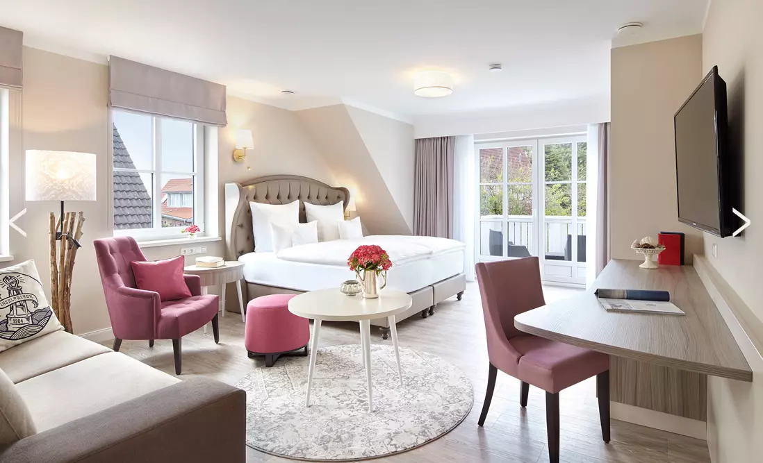 Header Zimmerpfeile Hell eingerichtetes Hotelzimmer mit Doppelbett, Sitzbereich, Schreibtisch und Balkon im Hotel Inselfriede auf Spiekeroog