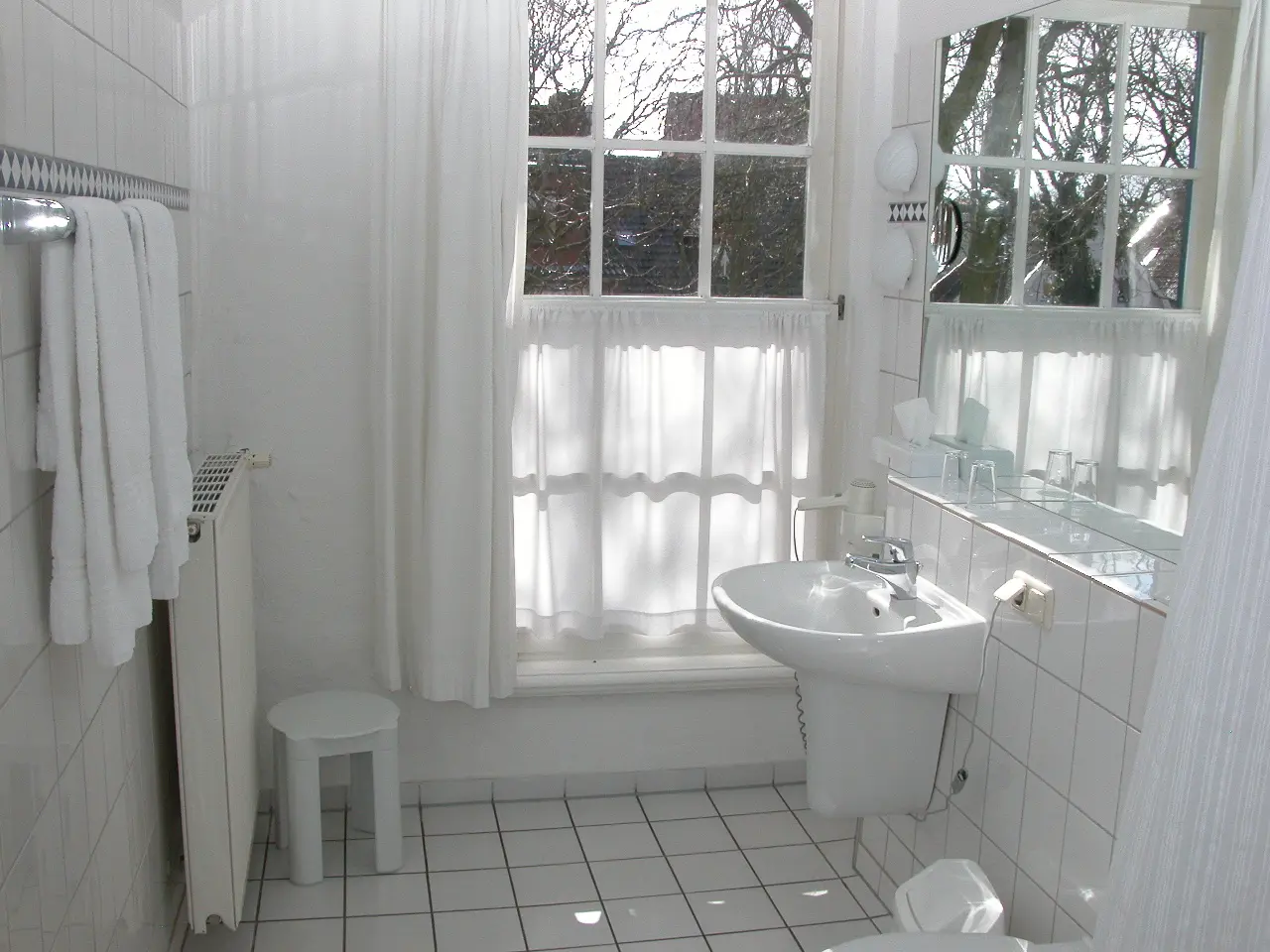Helles Badezimmer im Hohes Haus Greetsiel mit weißem Waschbecken, Handtüchern, großem Fenster und weiß gekachelten Wänden und Boden