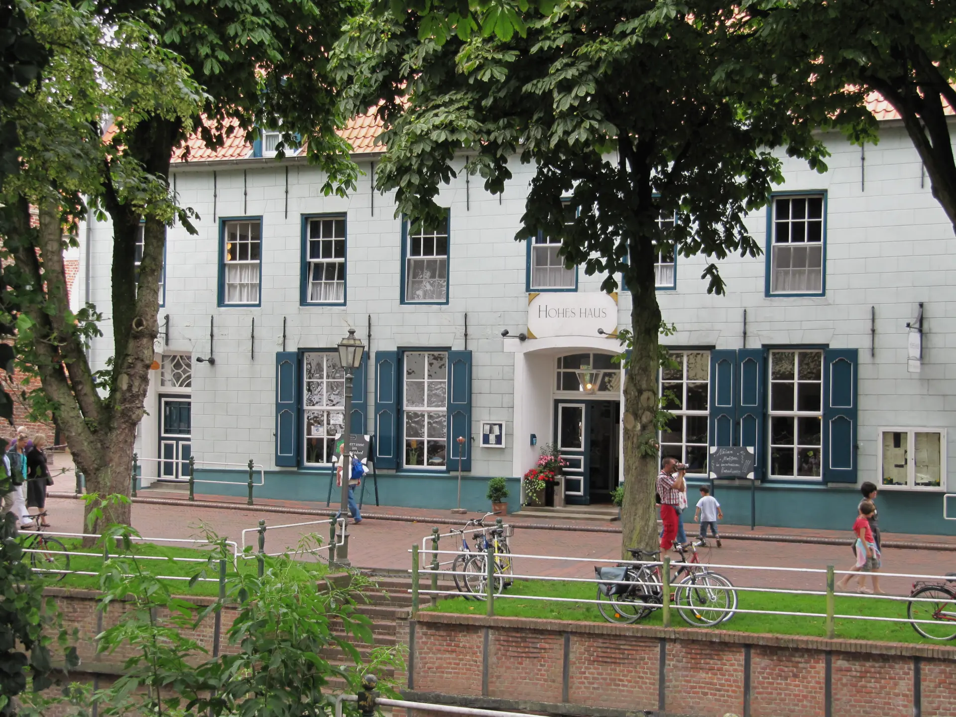 Fassade des historischen Hohes Haus in Greetsiel mit blauen Fensterläden, Bäumen und Fußgängern vor dem Gebäude