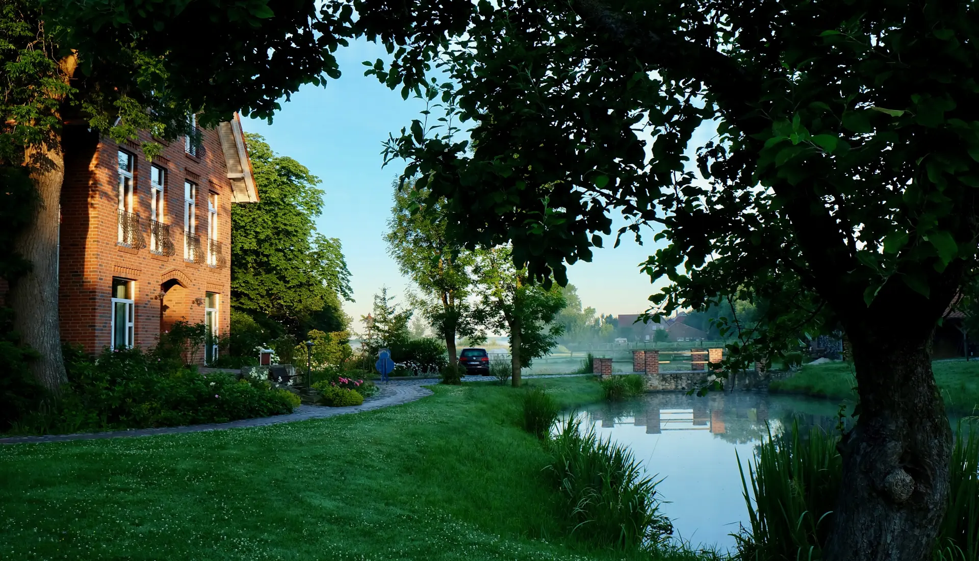 Hotel Kunze Hof Wattenmeer Jadebusen Außenansicht Seminarhotel Kunze-Hof am Wattenmeer mit rotem Backsteinhaus, Bäumen, grünem Rasen und ruhigem Wassergraben bei Sonnenaufgang