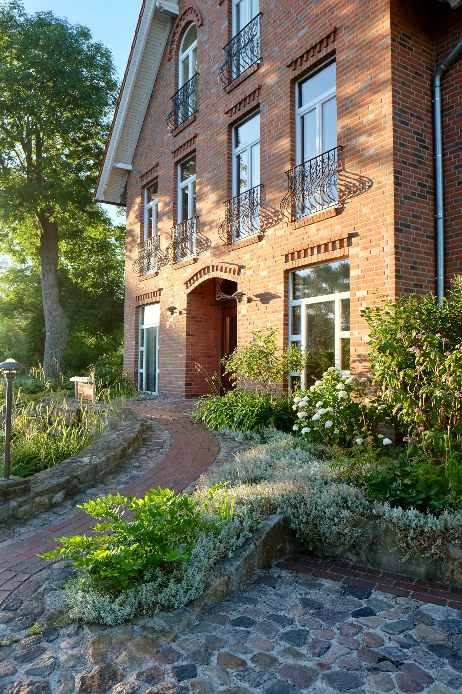 Hotel Kunze Hof Wattenmeer Jadebusen Außenansicht Backsteinhaus vom Seminarhotel Kunze-Hof in Wattenmeer mit Terrasse, Balkonen, gepflegtem Garten und Weg entlang der Fassade bei Sonnenlicht