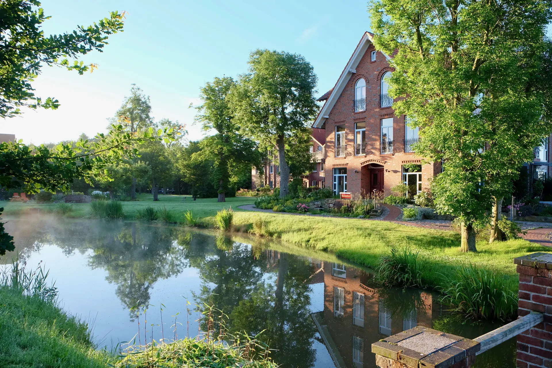 Hotel Kunze Hof Wattenmeer Jadebusen Außenansicht Seminarhotel Kunze-Hof aus rotem Backstein am Flussufer mit Bäumen und grüner Wiese im Wattenmeer bei klarem Himmel
