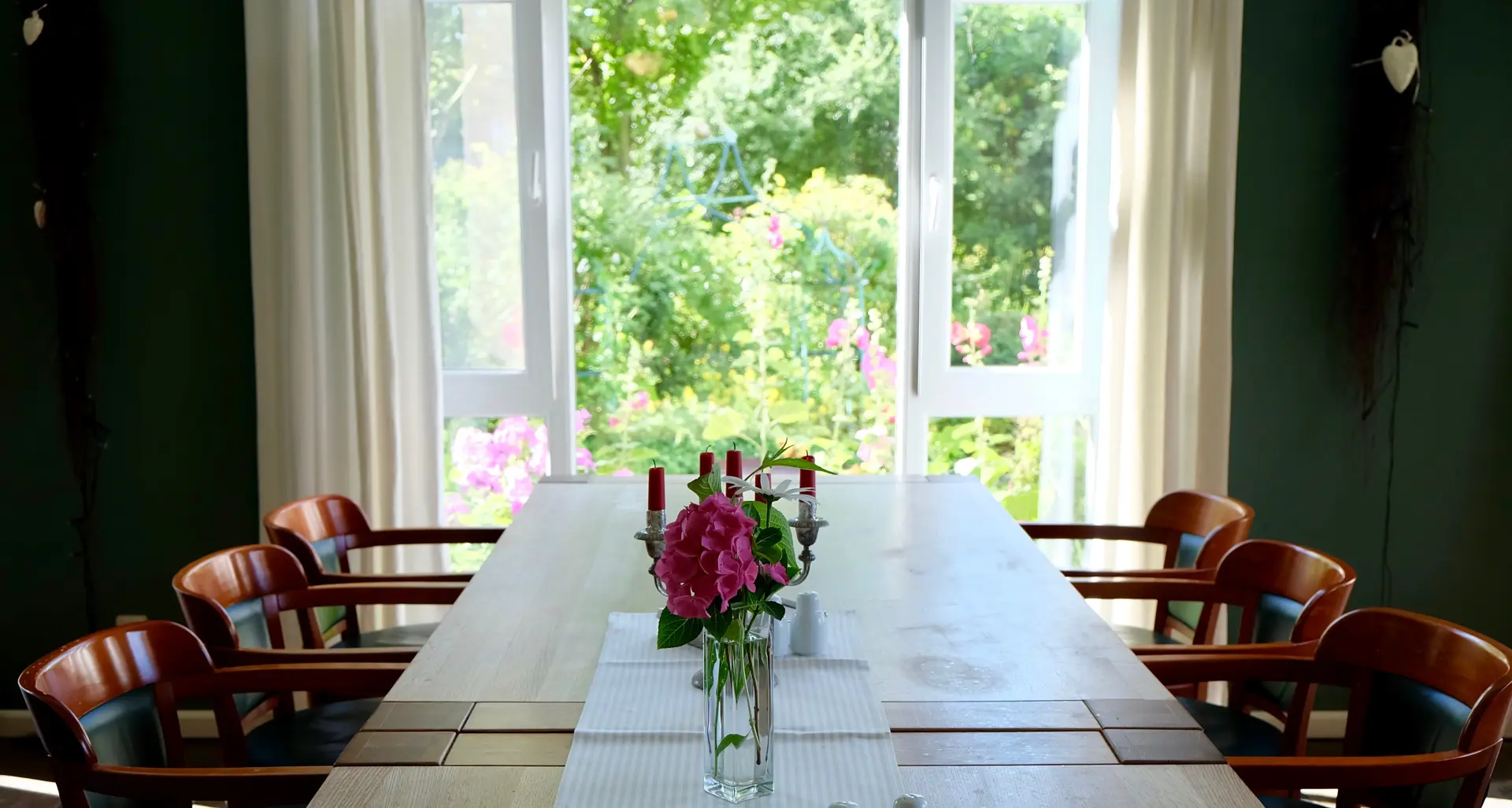 Hotel Kunze Hof Wattenmeer Jadebusen Tisch Großer Konferenztisch mit acht Stühlen, Kerzenständern und Blumen vor offenem Fenster mit Blick auf grünen Garten im Seminarhotel Kunze-Hof Wattenmeer