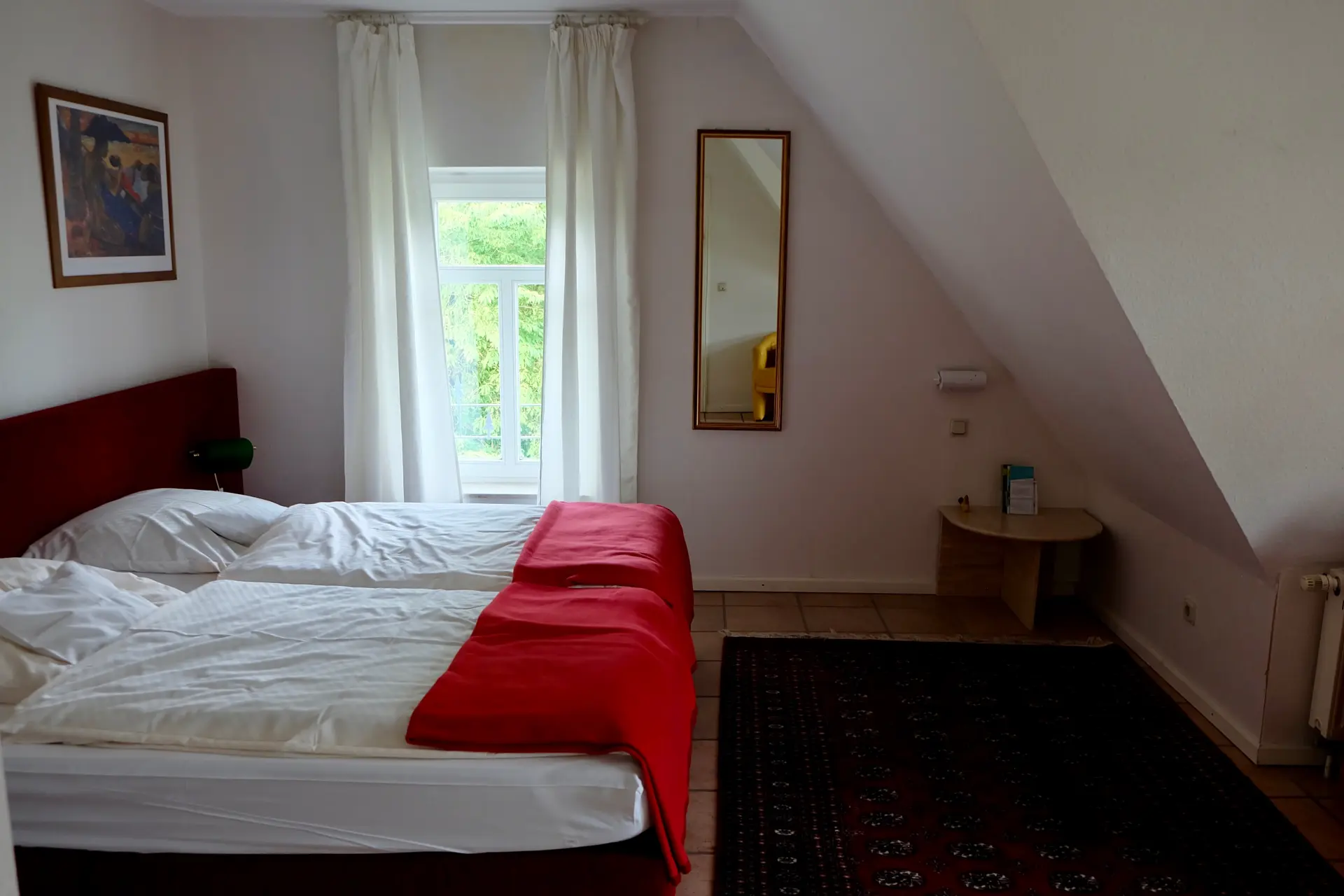 Zimmerbeispiel Gemütliches Hotelzimmer im Seminarhotel Kunze-Hof mit zwei Einzelbetten, roten Decken, Fenster mit Blick ins Grüne und schlicht eingerichteter Dachgeschosswohnung