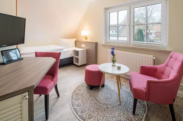 Hotel Inselfriede Spiekeroog Einzelzimmer 43 Hell eingerichtetes Hotelzimmer mit Einzelbett, Schreibtisch, Fernseher und gemütlicher Sitzecke mit rosa Sessel und kleinem Tisch in Hotel Inselfriede Spiekeroog