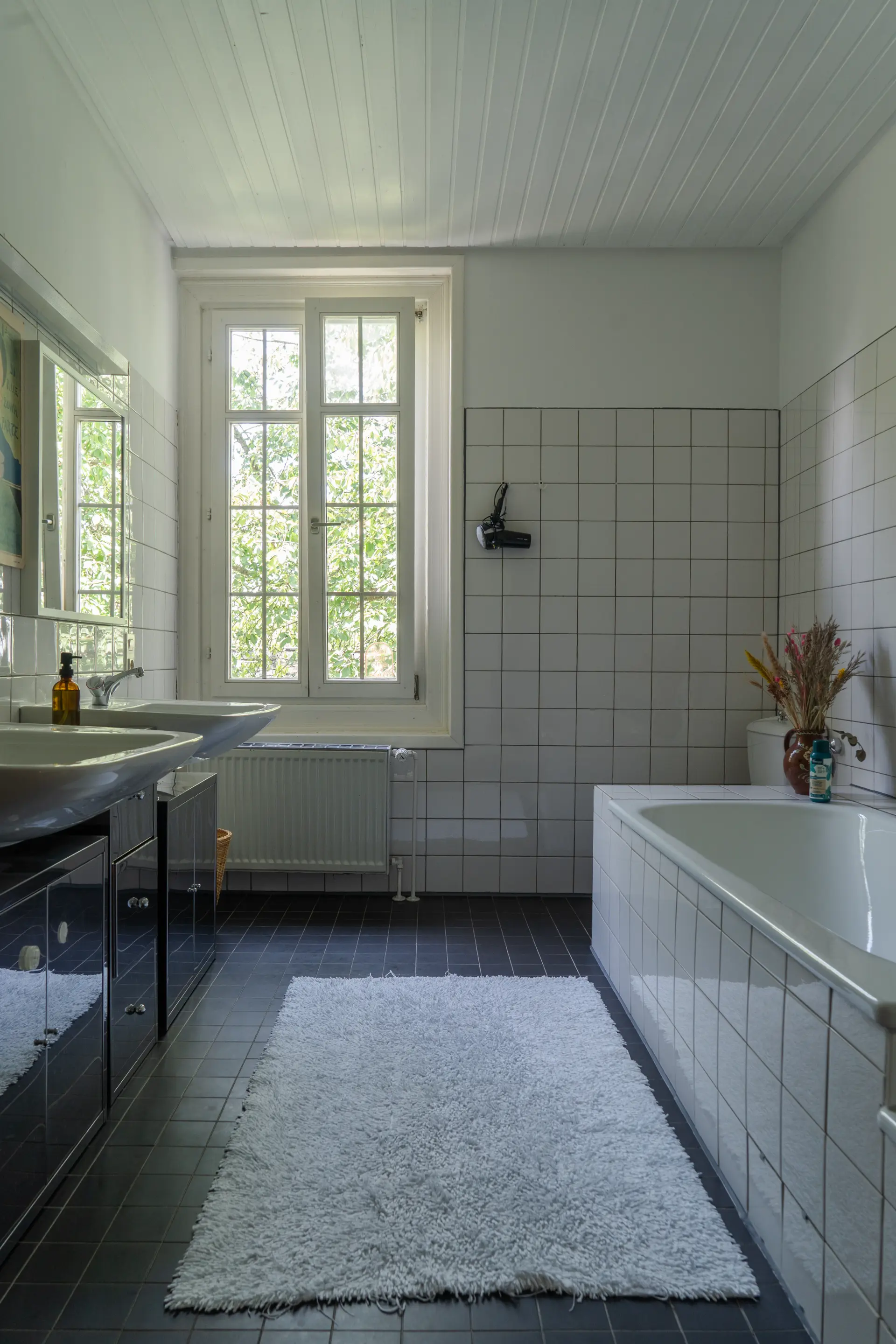 Helles Badezimmer im Landhaus Arnoth mit weißen Fliesen, Doppelwaschbecken, Badewanne, großem Fenster und flauschigem weißem Teppich in der Moselregion