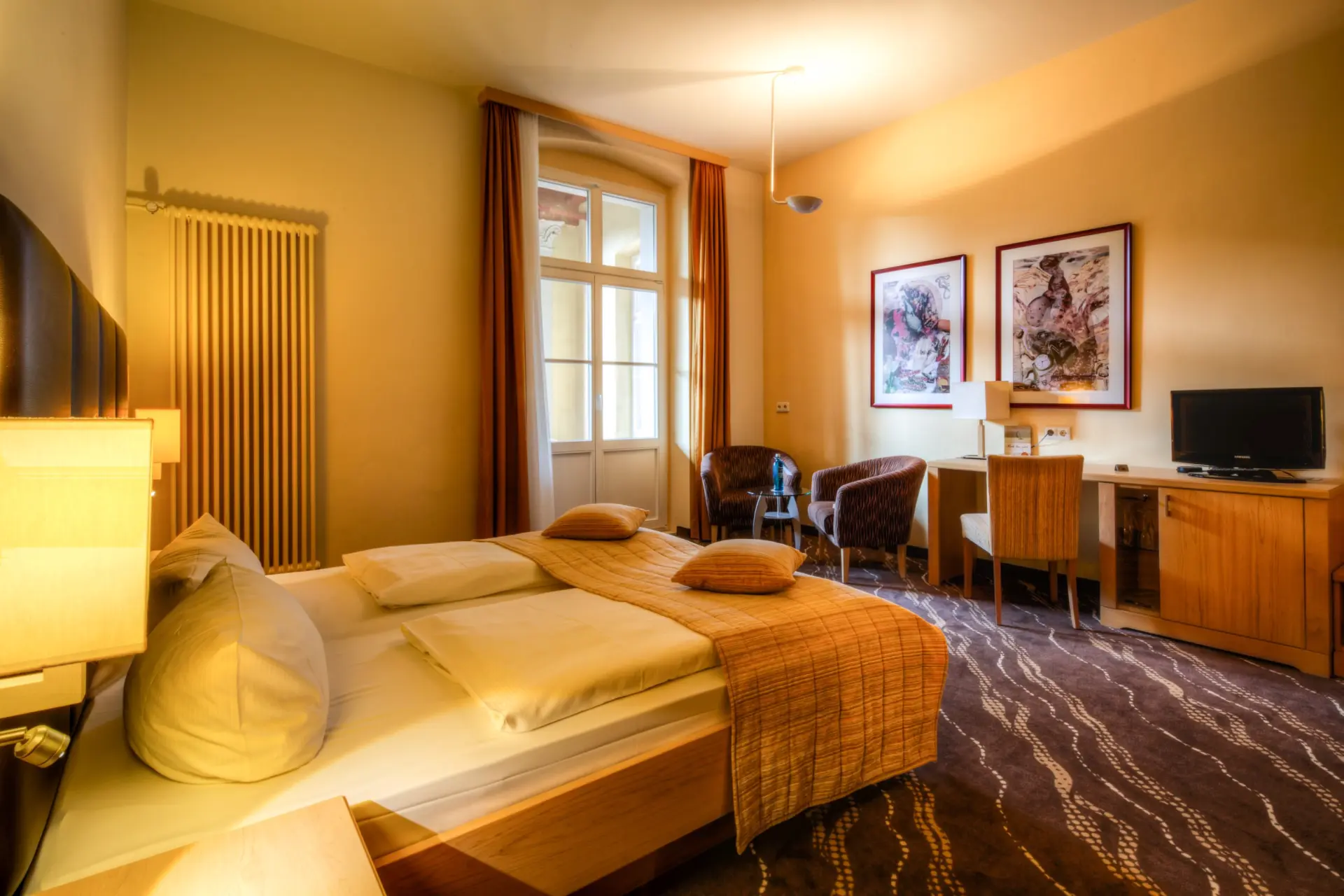 Hell eingerichtetes Hotelzimmer im Parkhotel Bad Schandau mit großem Bett, Schreibtisch, TV und zwei Sesseln vor Fenster mit Vorhängen