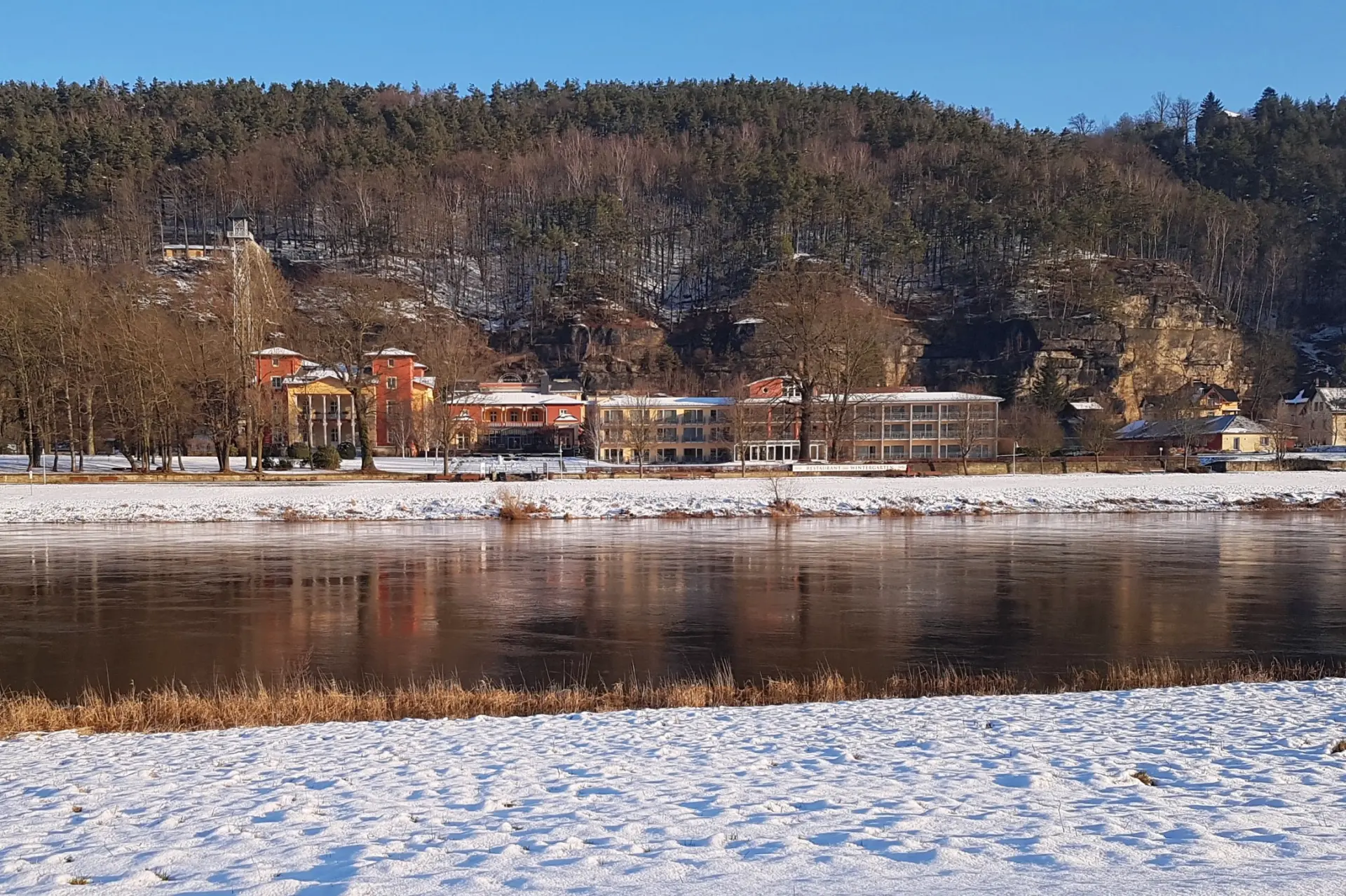 Parkhotel Bad Schandau mit umliegendem Schnee, Elbufer und bewaldeten Felsen in der winterlichen Sächsischen Schweiz
