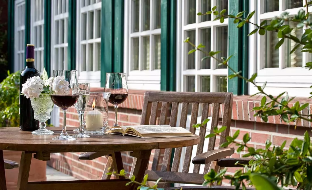 SL Terasse Holztisch mit Weinglas, Kerze, Blumen und Buch vor Fensterreihe eines Hotels im Außenbereich der Unterkunft Hotel Inselfriede auf Spiekeroog