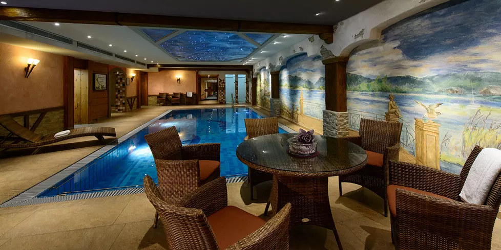 Indoor-Poolbereich im Steinbach-Hotel Berchtesgadener Land mit Korbsesseln, Tisch und Wandmalerei im gemütlichen Ambiente