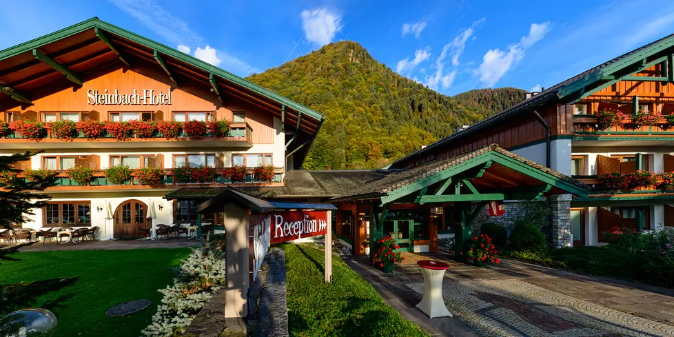 Vier Sterne Steinbach Hotel Ruhpolding Chiemgau Bayern Alpen Winterurlaub Sommerurlaub Ski Langlauf Biathlon Eingang Westernberg Bergpanorama Traditionelles Steinbach-Hotel in Berchtesgadener Land mit Alpenpanorama, blühenden Balkonkästen und Empfangsbereich im Sommer