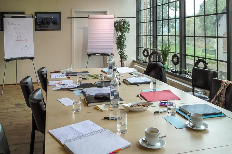 Konferenztisch im Hotel Birschel-Mühle mit Ordnern, Notizblöcken, Wasserflaschen, Kaffeetassen und Flipcharts vor großen Fenstern mit Blick ins Grüne