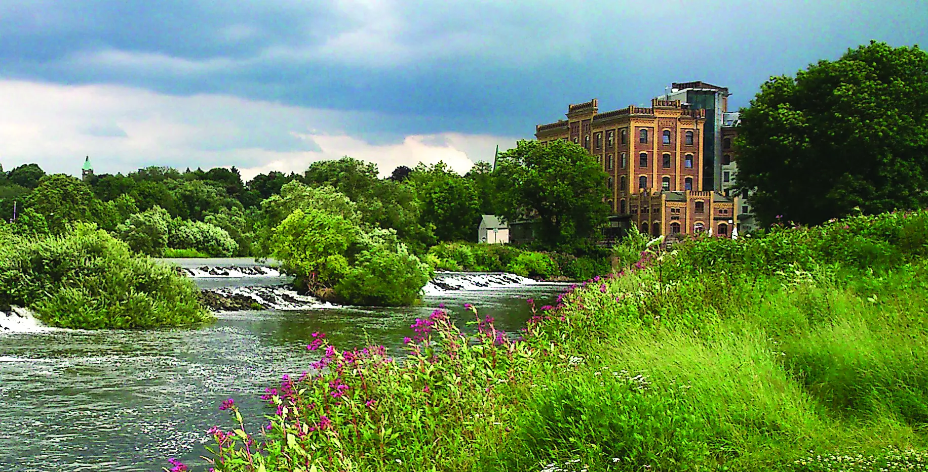 Fluss mit kleinen Wasserfällen und grüner Ufervegetation vor dem historischen Backsteingebäude des Hotel Birschel-Mühle in Hattingen