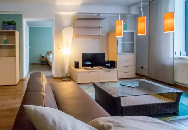 Modernes Wohnzimmer mit braunem Sofa, Glas-Couchtisch, TV, Stehlampe und orangen Hängelampen im Hotel Birschel-Mühle in Hattingen