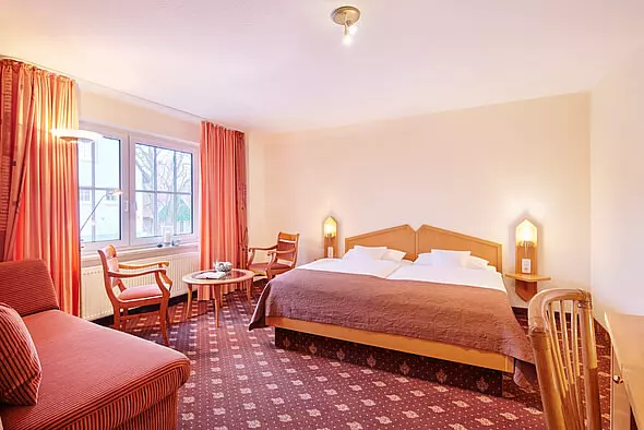 Hotelzimmer mit Doppelbett, zwei Sesseln, Sofa, Teppichboden und großen Fenstern im Hotel Inselfriede auf Spiekeroog