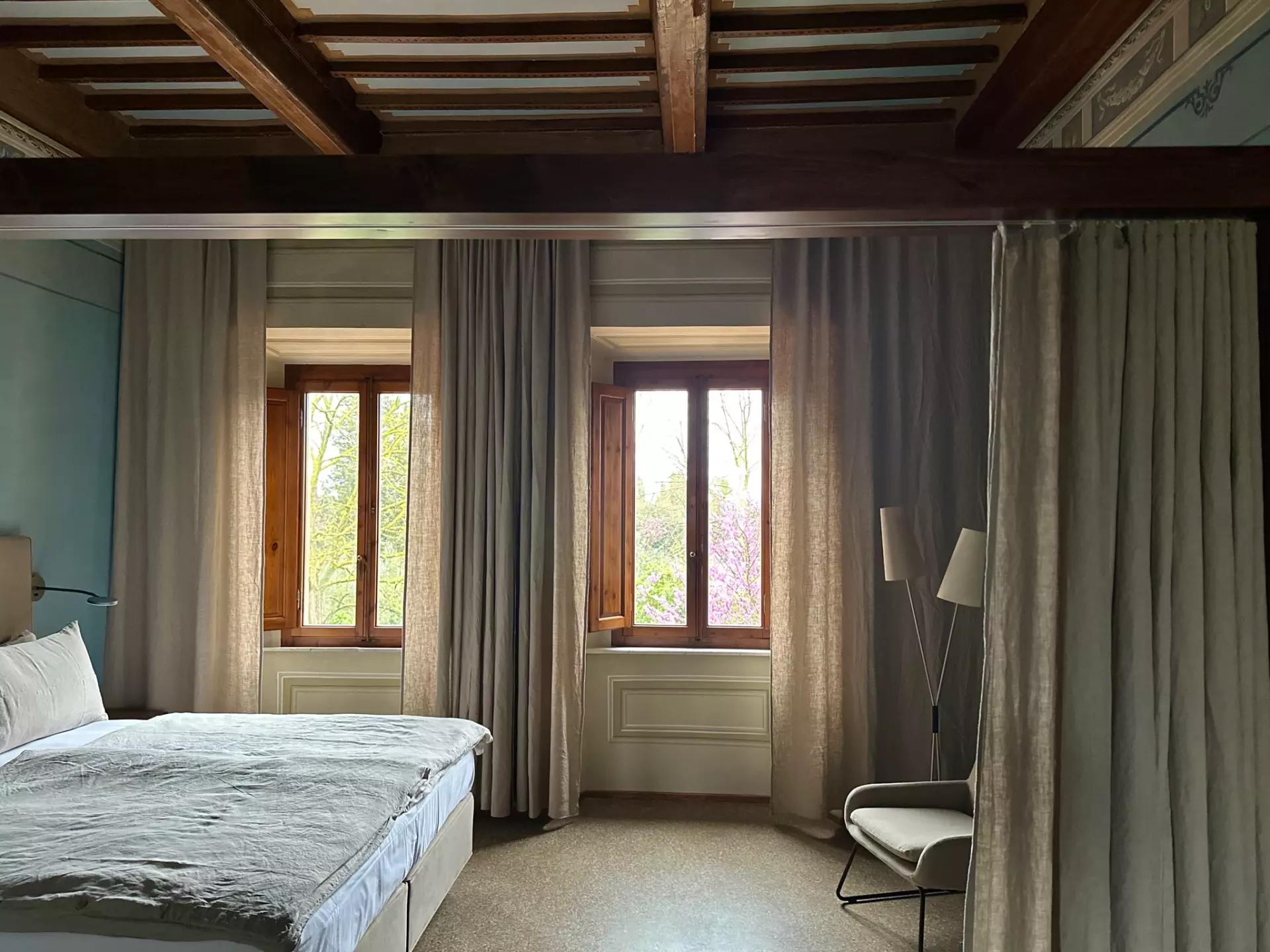 Doppelzimmer Ansicht 2 Schlafzimmer mit großen Fenstern, Vorhängen, Holzbalkendecke, grauer Bettdecke und gemütlichem Sessel in einer Unterkunft in der Toskana