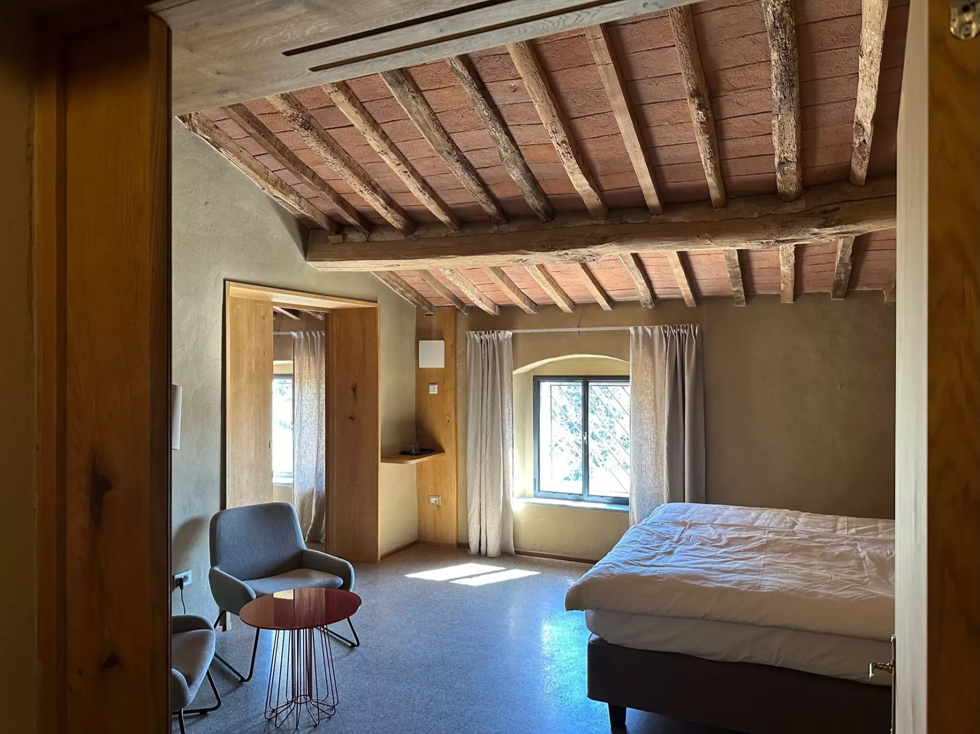 Helles Schlafzimmer in Toskana-Unterkunft mit rustikalem Holzbalkendecken, Doppelbett, Fenster mit Vorhängen und gemütlicher Sitzecke mit Stühlen und kleinem Tisch