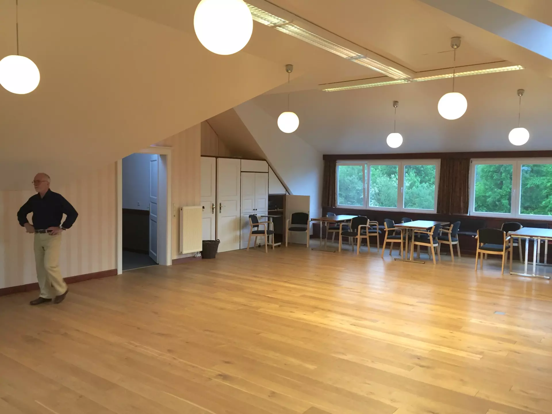 Großer heller Raum mit Holzboden, weißen Wänden, mehreren Tischgruppen am Fenster und hängenden Kugelleuchten im Atelier im Hotel Wattenmeer