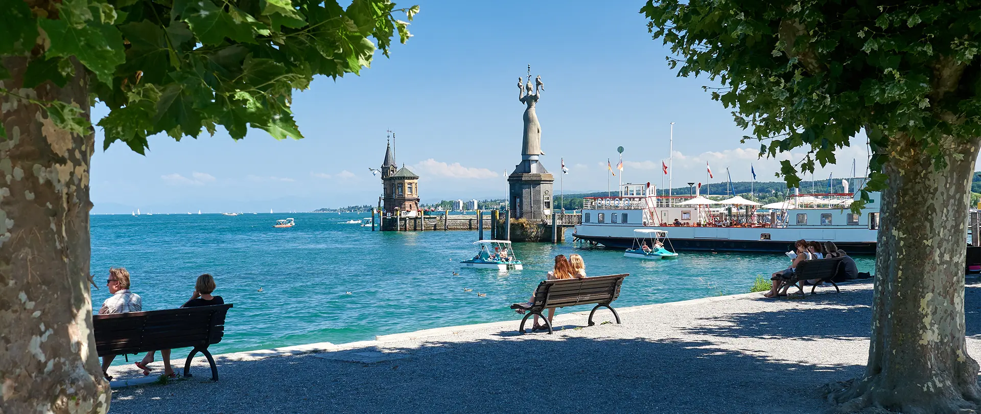 Blick auf den Bodensee in Konstanz mit Imperia-Statue, Hafen, Sitzbänken und Menschen unter schattigen Bäumen bei klarem Himmel