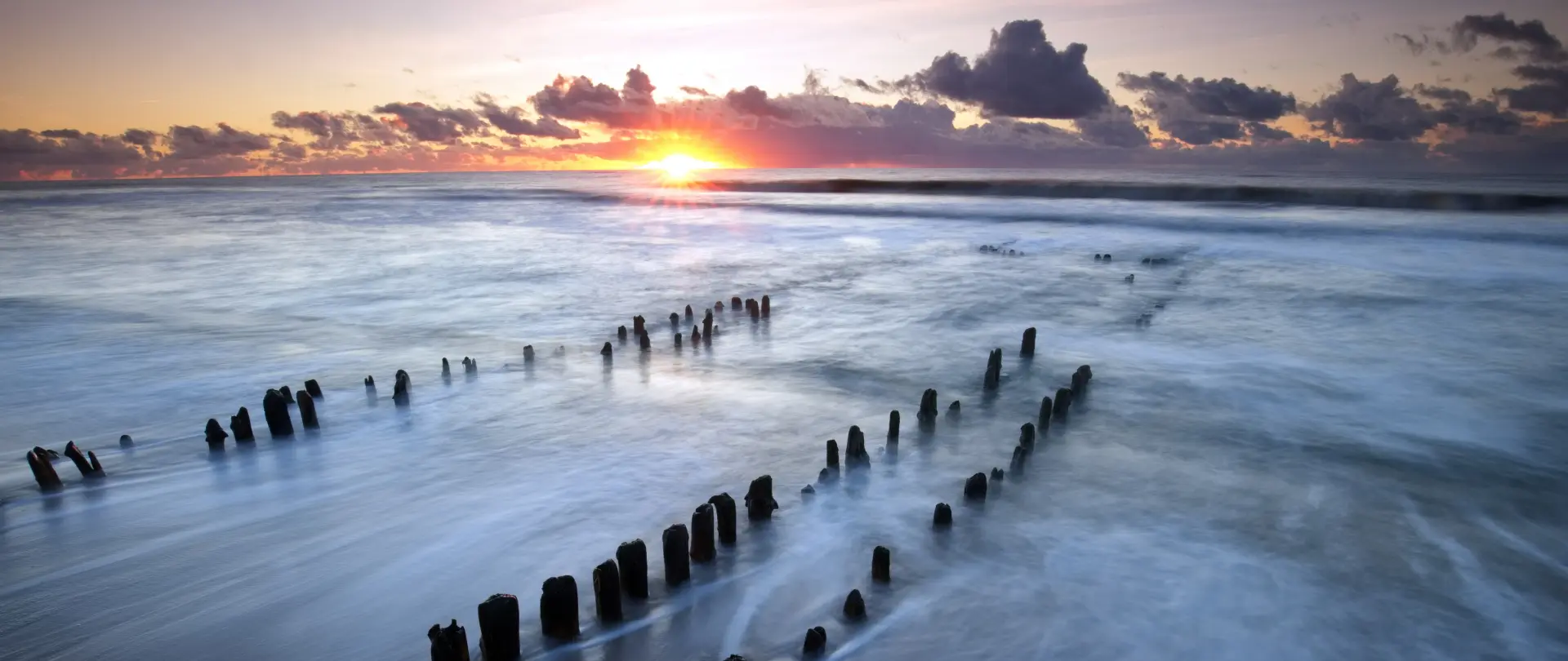Sonnenaufgang über ruhigem Meer mit Holzpfählen im Wasser, natürliche Stimmung und Langzeitbelichtung von Marko Sarcevic