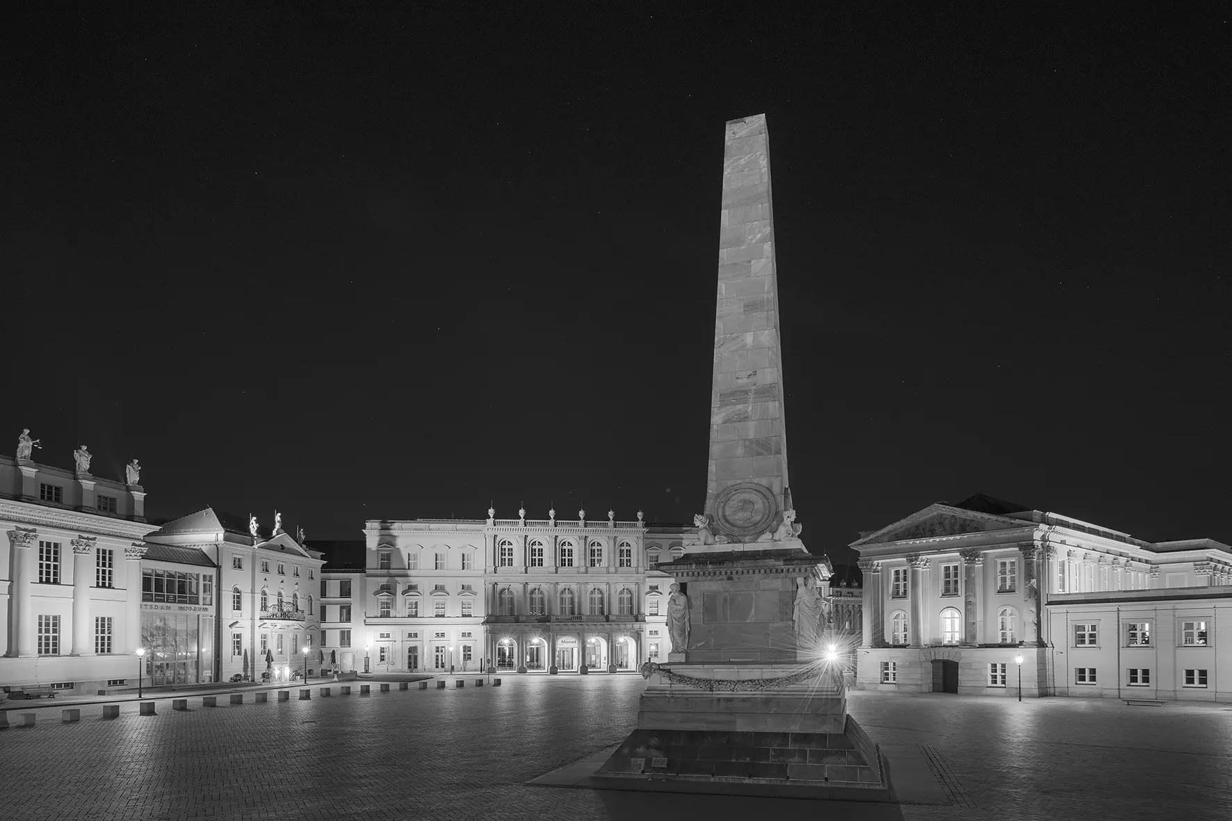 Schwarz-weiße Stadtaufnahme bei Nacht mit beleuchtetem Obelisken und Gebäuden, klare Licht- und Schattenkontraste von Jutta Engelage