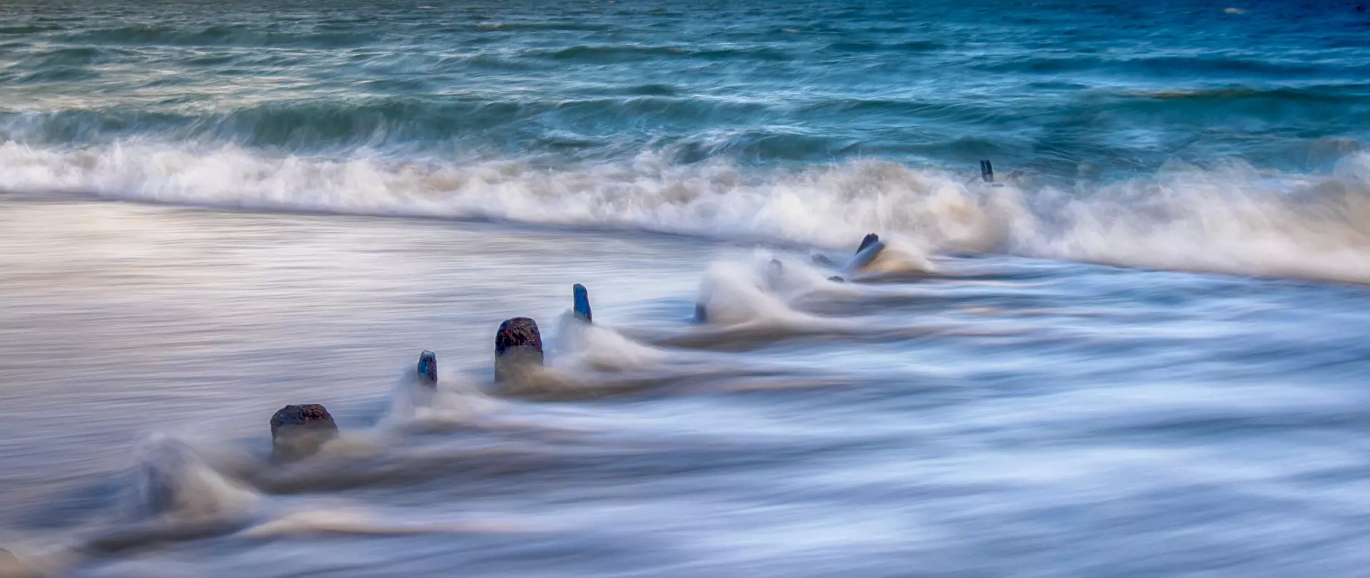 Langzeitbelichtete Meereswellen umspülen Holzpfähle in sanften Blautönen, abstrahiert von Marion Hogl mit besonderem Fokus auf Licht und Farbe