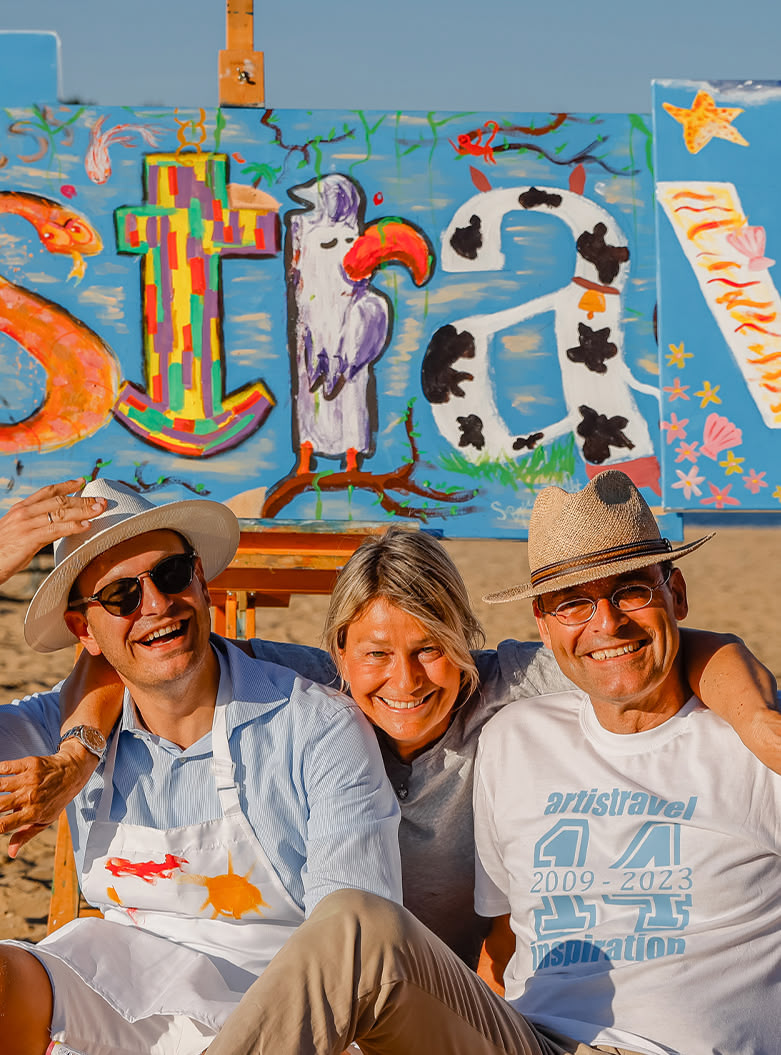 Tom, Dani und Sebastian Gothe sitzen im Sand vor 3 Leinwänden mit gemaltem artistravel-Logo