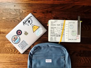 Bullet Journal - Habit Tracker / Malen und Zeichnen lernen. Foto: Matt Ragland/Unsplash Ein Rucksack, ein MacBook mit Stickern und ein offenes Notizbuch liegen auf einem Tisch