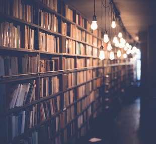 Creative Writing/Kreatives Schreiben. Schreibkurs - Subgenres. Foto:Janko Ferlic/Unsplash Ein großes Bücherregal, das spärlich von herabhängenden Lampen spärlich beleuchtet wird