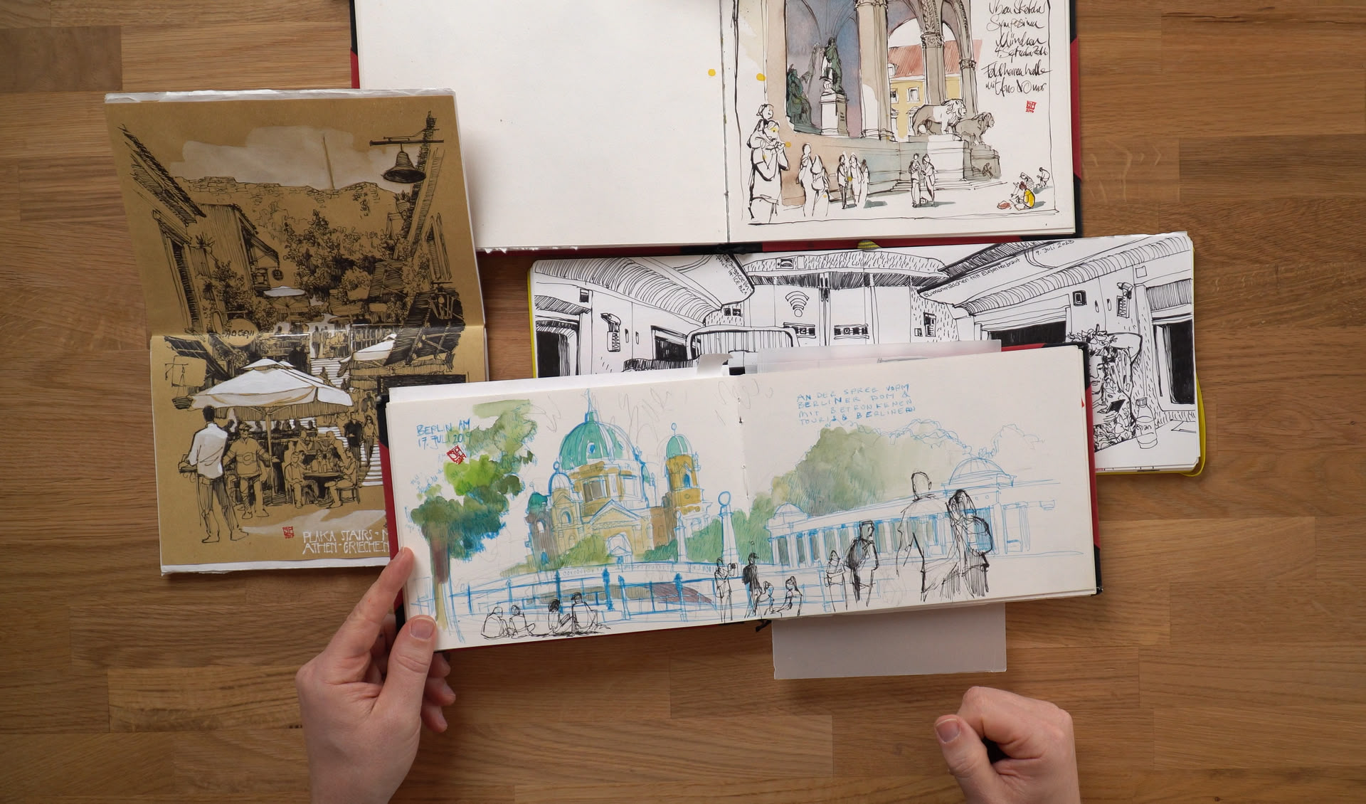 Blick aus der Vogelperspektive auf einen Tisch mit Skizzenbüchern und den Händen von einer Person, die die Sketches darin zeigt