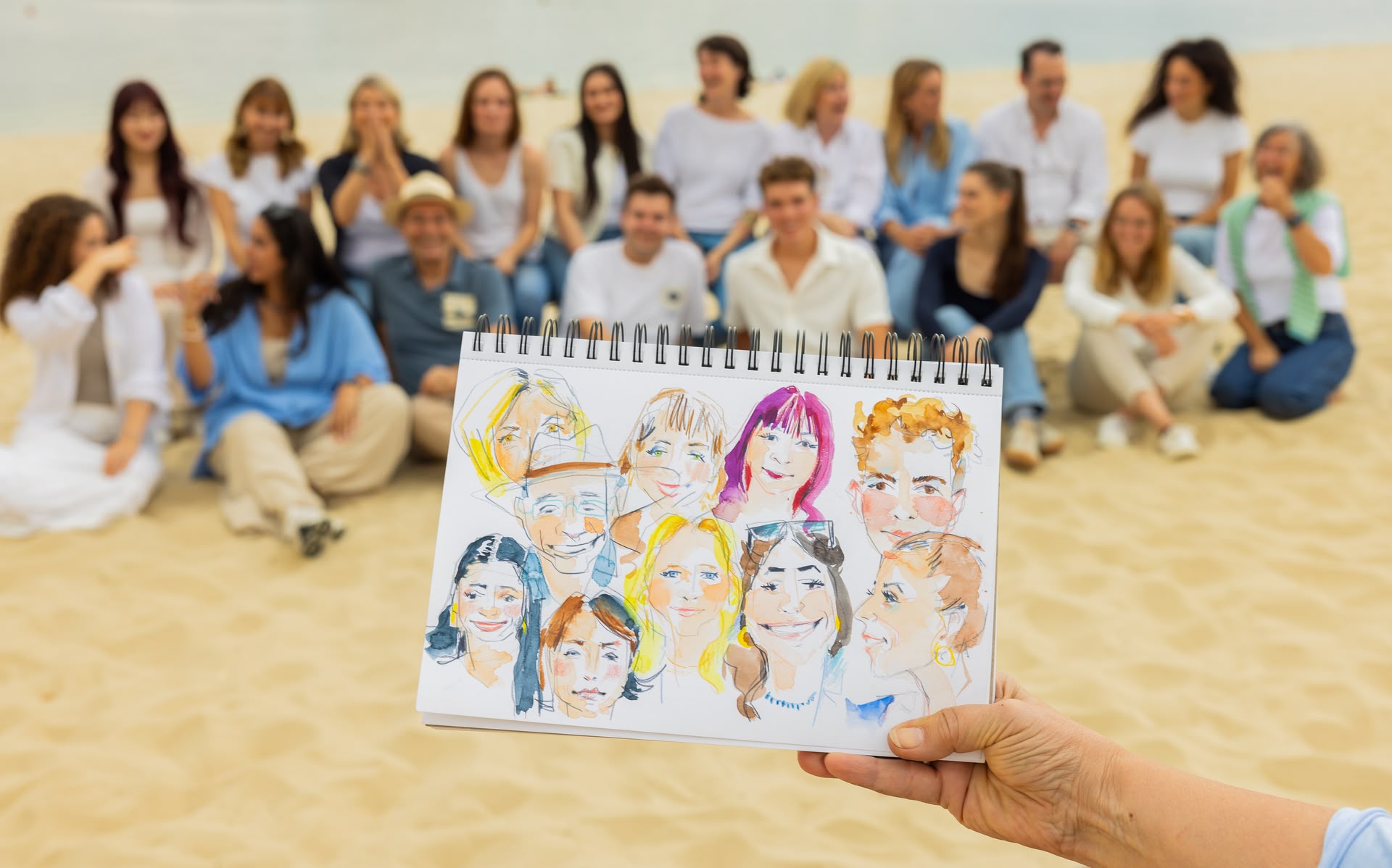 Ein Sketch von einer Gruppe Menschen wird am Strand vor die Menschengruppe (das artistravel-Team) gehalten, die das Motiv für das Werk war