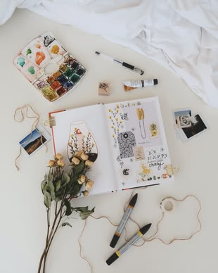 Estee Janssens (Unsplash) Rund um ein offenes Notizbuch liegen verschiedene Dinge (Stifte, Farbkasten, Rosen, Fotos)